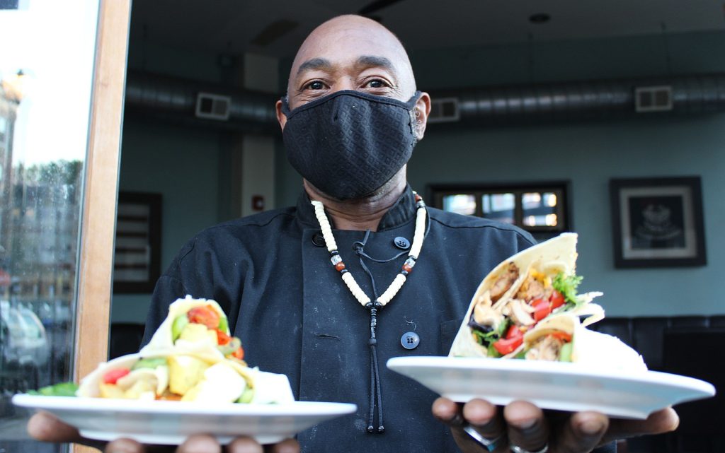 Photo of Chef in black chef coat holding two plates with wraps on them towards the camera.